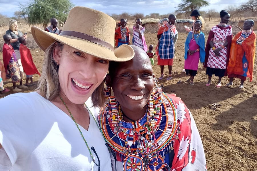Una mujer con sombrero de paja posa junto a un auténtico guerrero masai mientras otros conversan de pie en el fondo | Go2Africa