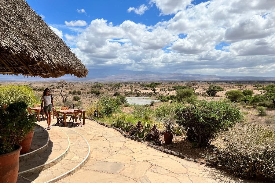 A woman walks towards the camera around the curve of a thatched building with the plains of Amboseli to the right of the frame | Go2Africa