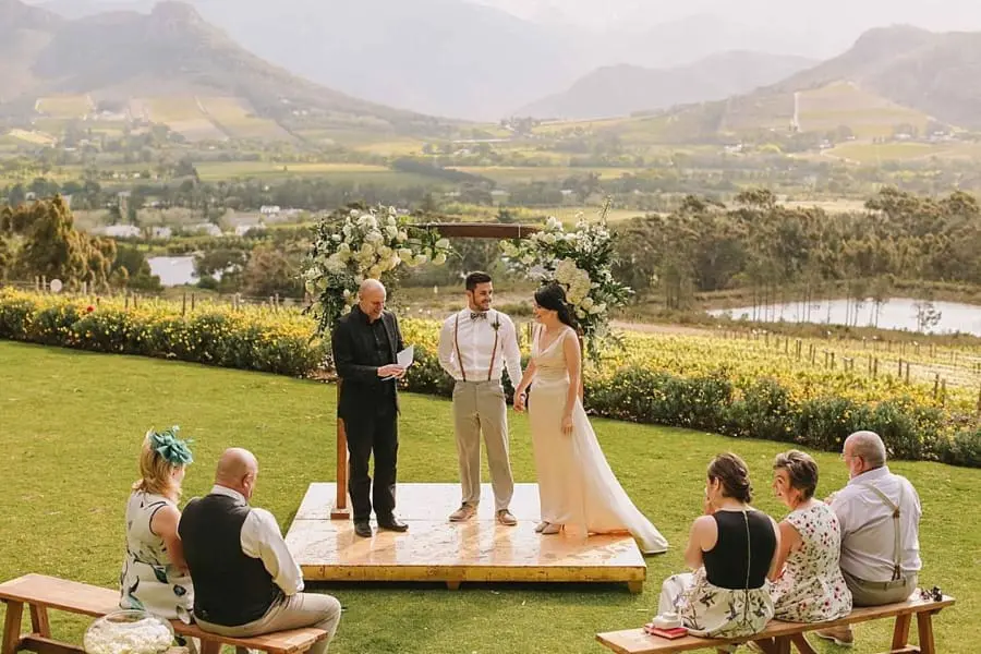 Una pareja de pie en una plataforma frente a su familia y amigos casándose con las montañas de Franschhoek como telón de fondo | Go2Africa