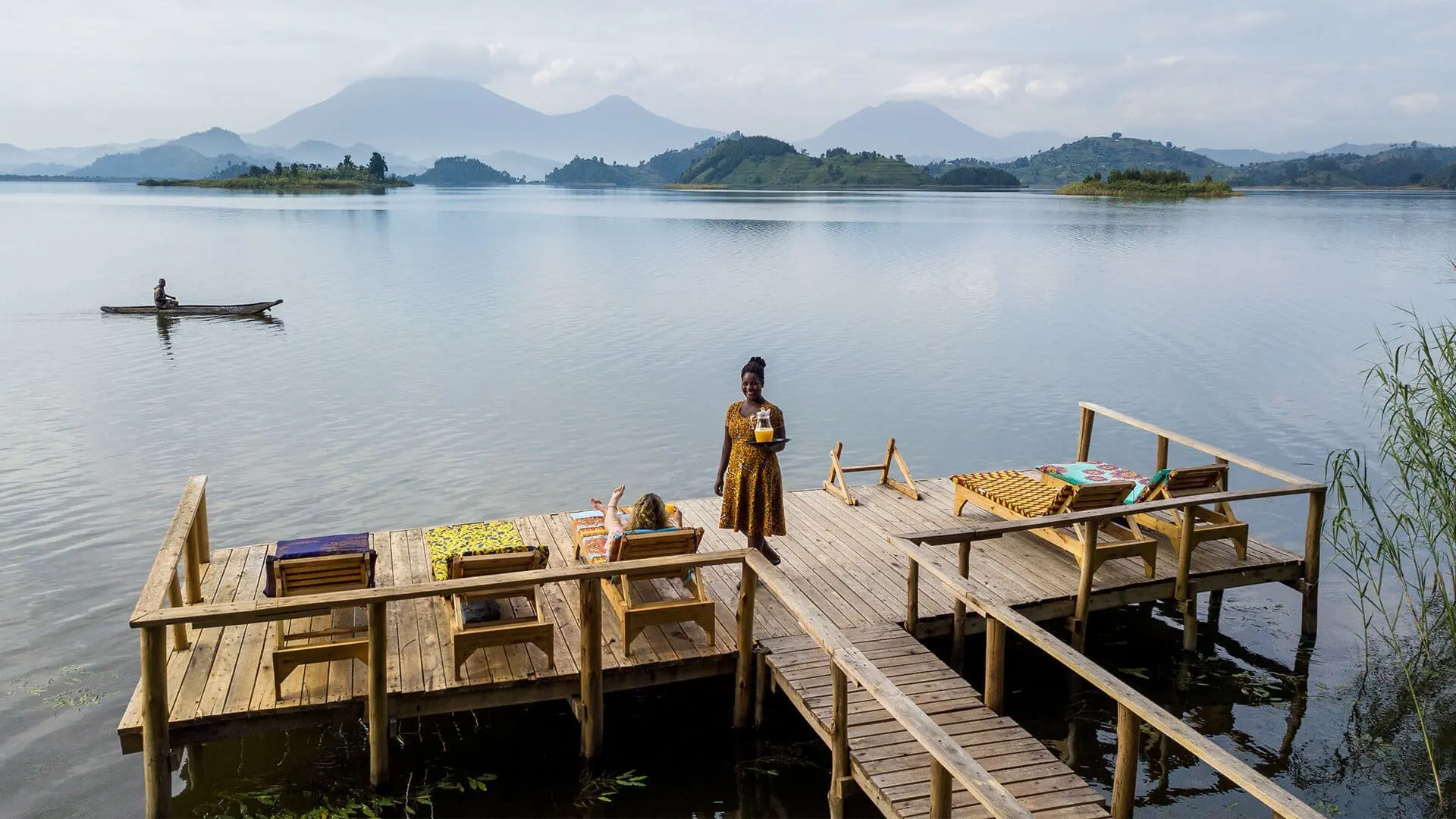 A woman sits on a lounger on a dock facing away from the camera while another person delivers a drink to her. Further in the distance a person canoes on the lake | Go2Africa