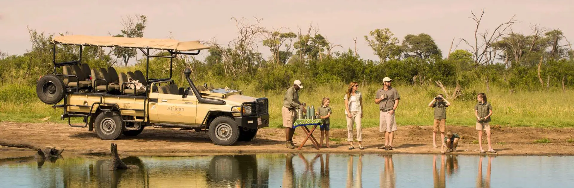 Una familia disfruta de una copa al atardecer con su guía junto a una masa de agua en primer plano | Go2Africa
