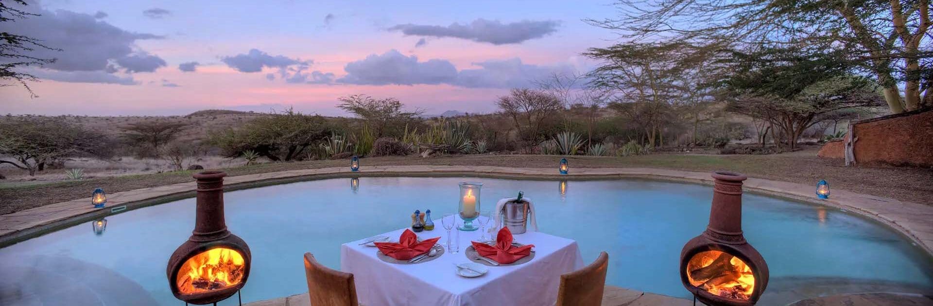 Una mesa para dos con vistas a la piscina en el Lewa Safari Camp acompañada de linternas y dos pequeños fuegos contenidos al atardecer | Go2Africa
