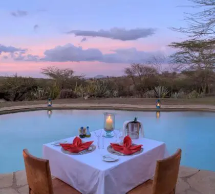 Una mesa para dos con vistas a la piscina en el Lewa Safari Camp acompañada de linternas y dos pequeños fuegos contenidos al atardecer | Go2Africa