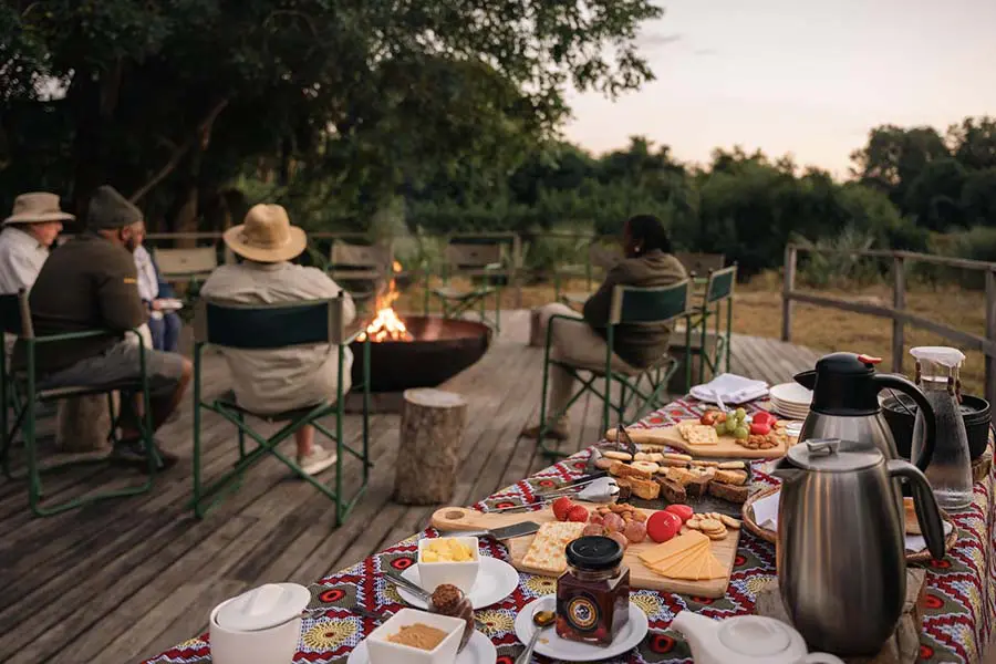 A table of snacks and food sits in the foreground while guests and a guide sit around a fire further back | Go2Africa