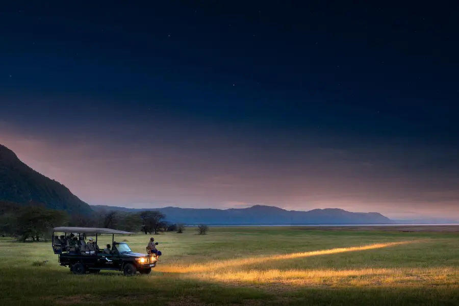 Open safari vehicle on a night drive in Lake Manyara, with headlights casting a beam across grassy plains under a star-filled twilight sky | Go2Africa